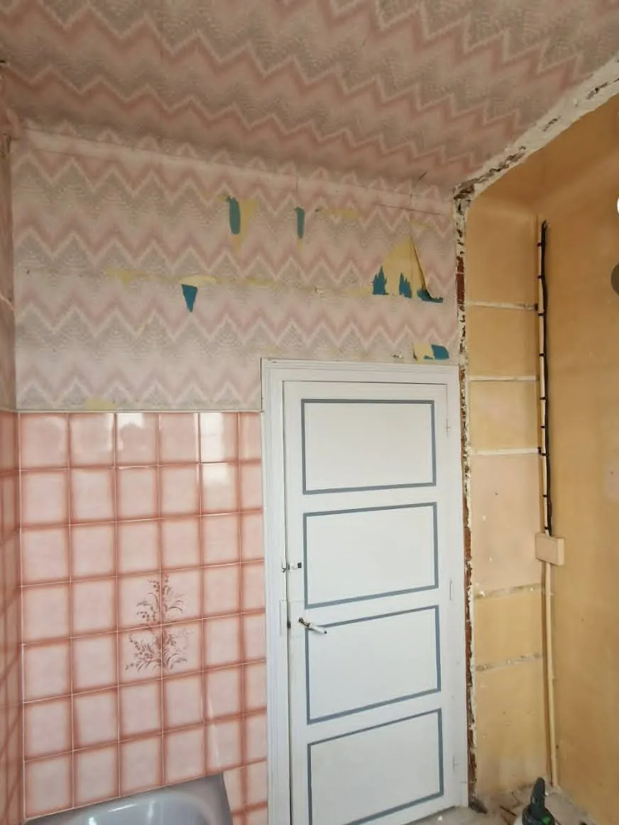 Salle de bain — côté porte avant travaux : Papier peint à chevrons, carrelage rose et huisserie vernie.